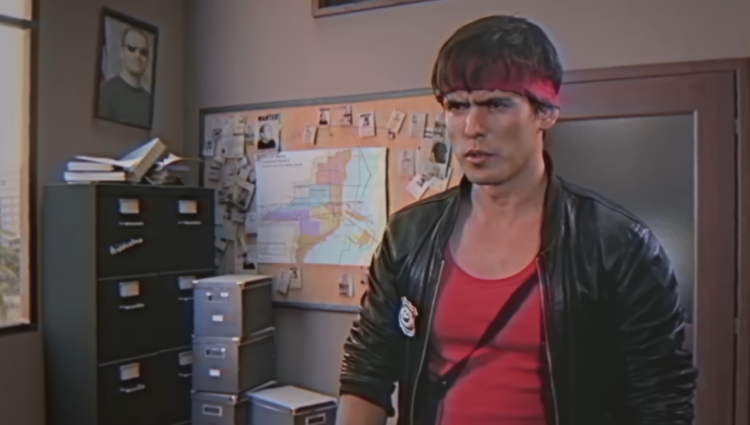 Kung Fury, a US detective dressed in a black leather jacket with a red vest and matching headband. He is in a police captain's office with filing cabinets and a pinboard behind him.